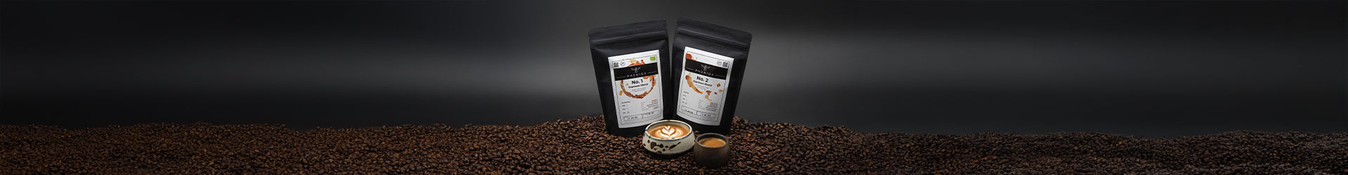 PHENIOX Kaffeeröstungen No. 1 und No. 2 sowie eine PHENIOX Cappuccino Tasse Barista und eine PHENIOX Espressotasse Timeout stehen in einem Berg voll Kaffeebohnen vor einem schwarzen Hintergrund.